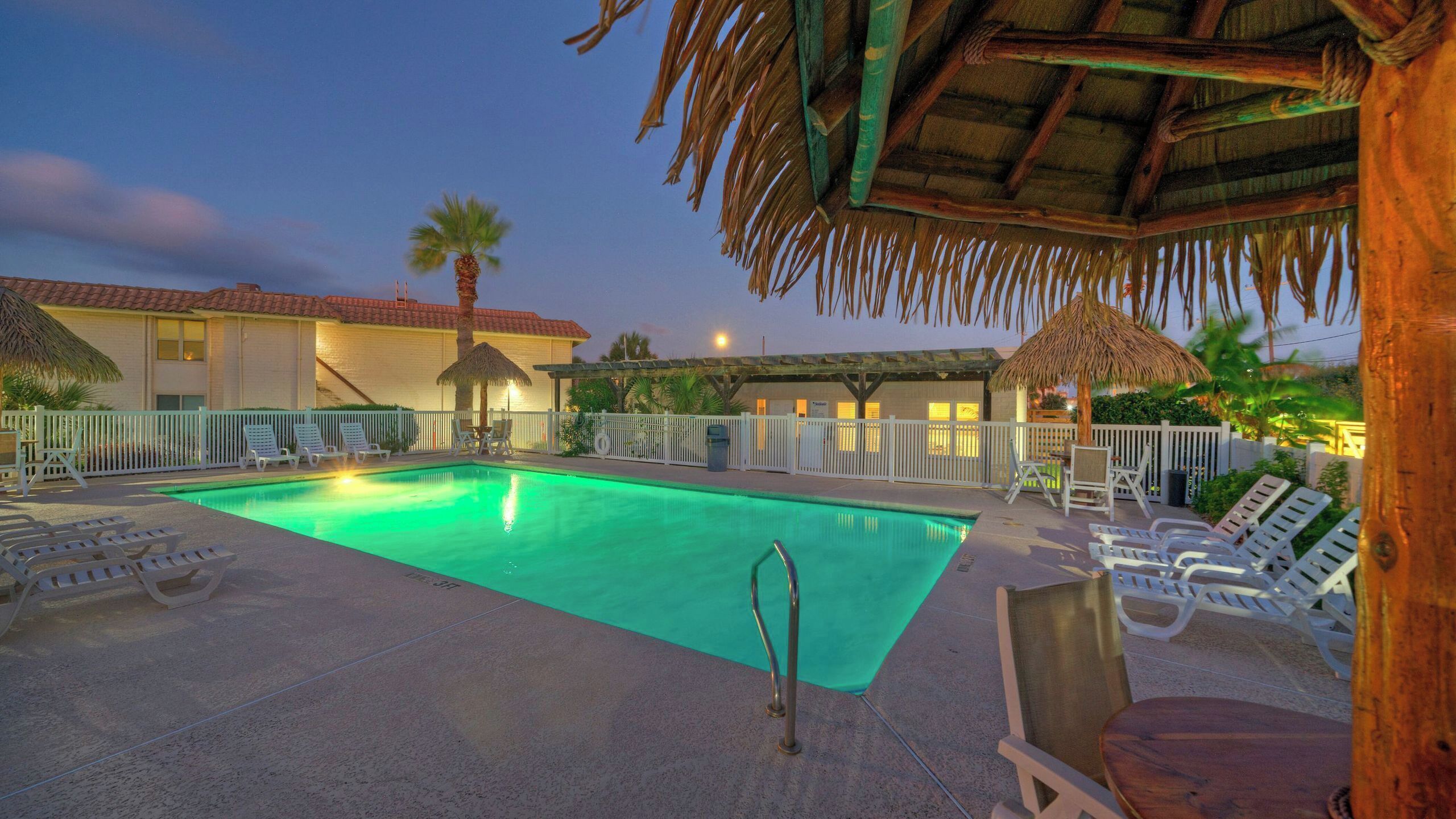 A swimming pool with lounge chairs, a thatched roof, and a white fence.