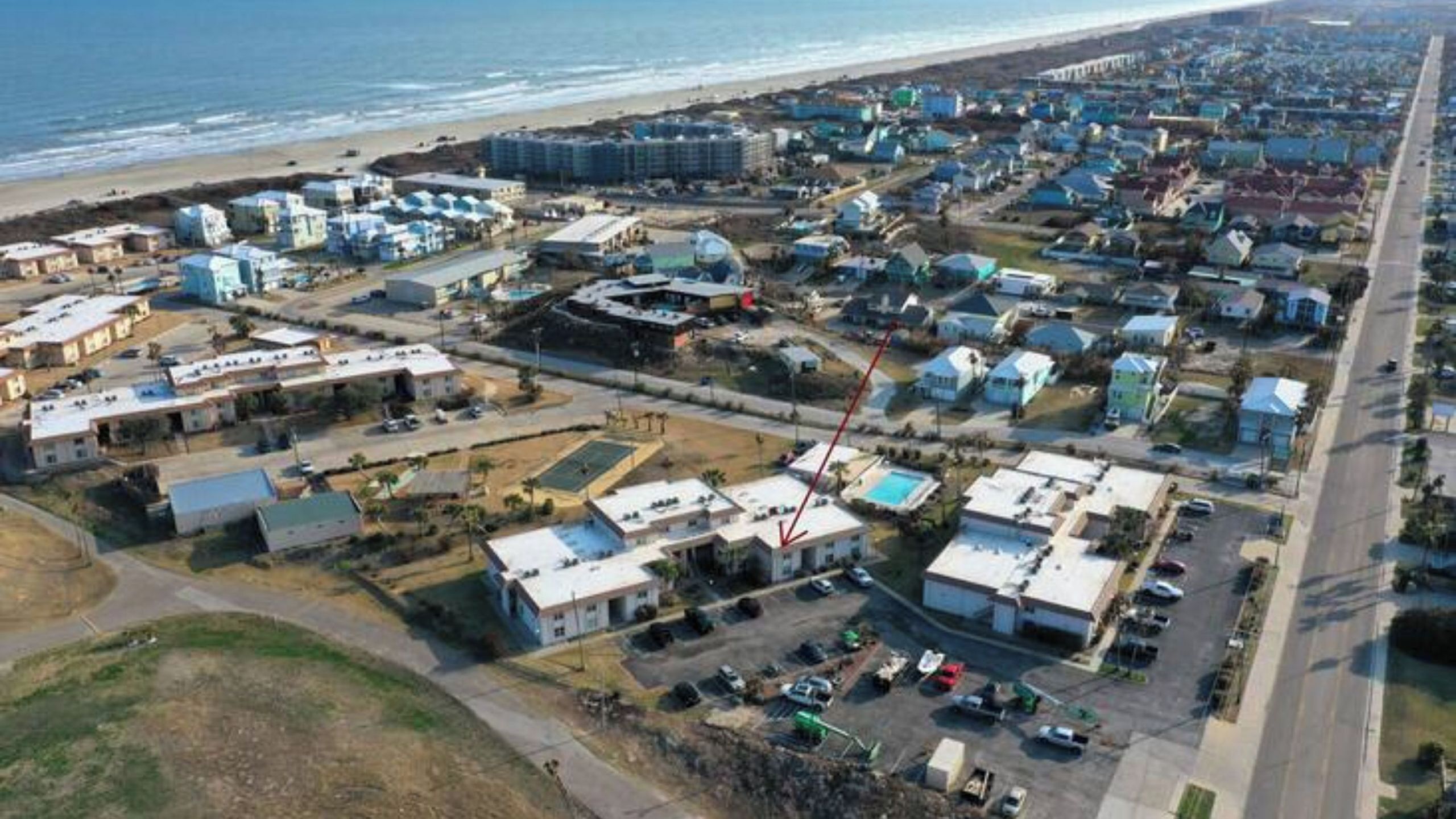 An aerial view of a coastal residential area with multiple buildings, a beach, and a road.