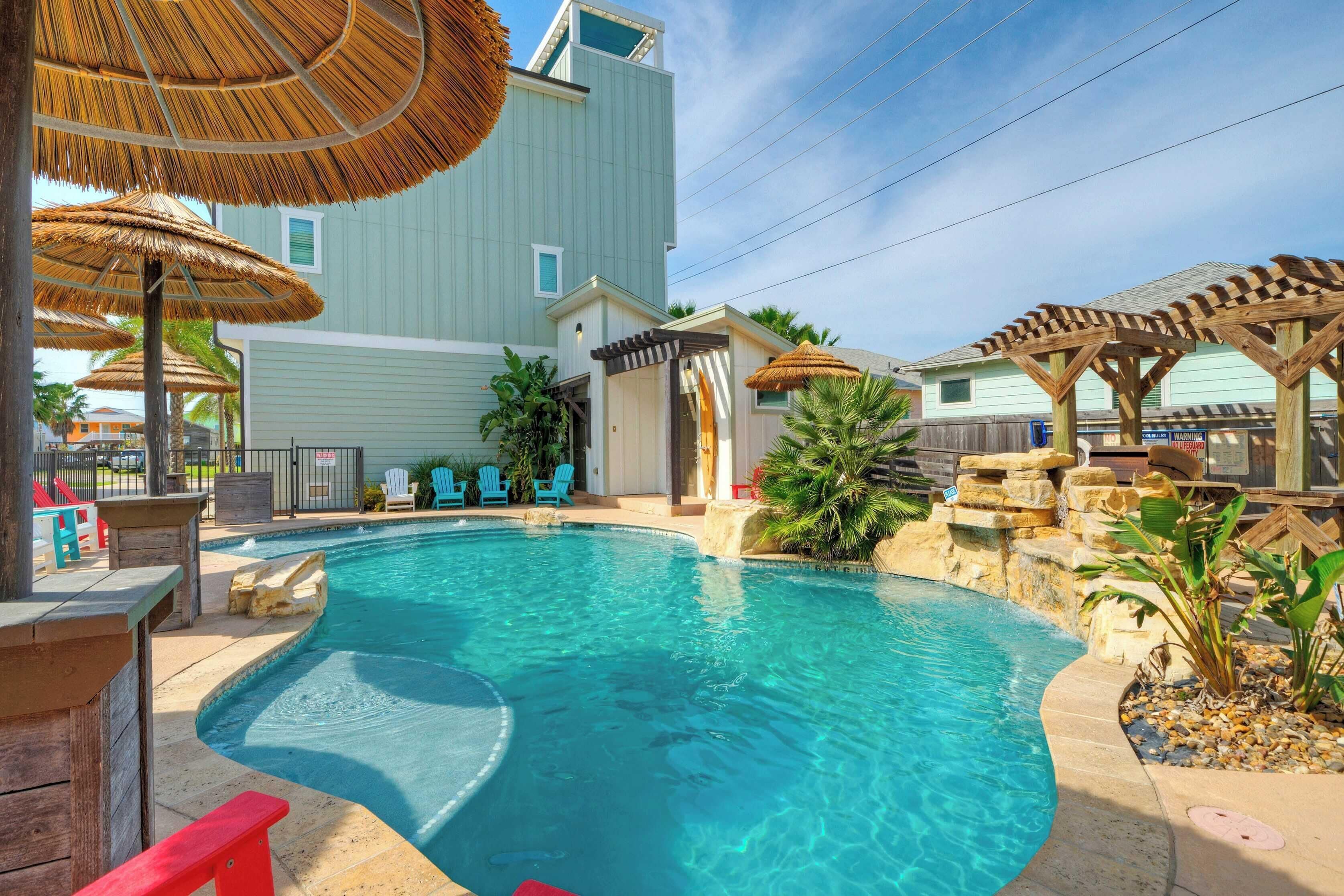 A swimming pool with a curved edge, surrounded by a wooden deck, a pergola, and thatched umbrellas.