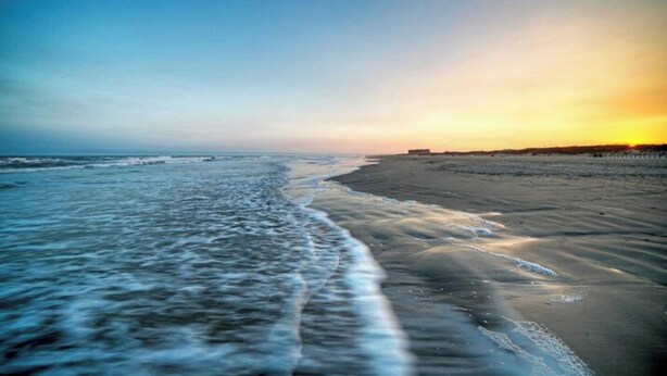 Ein Strand mit sanft anschwellenden Wellen, einem klaren Himmel und einem fernen Horizont.