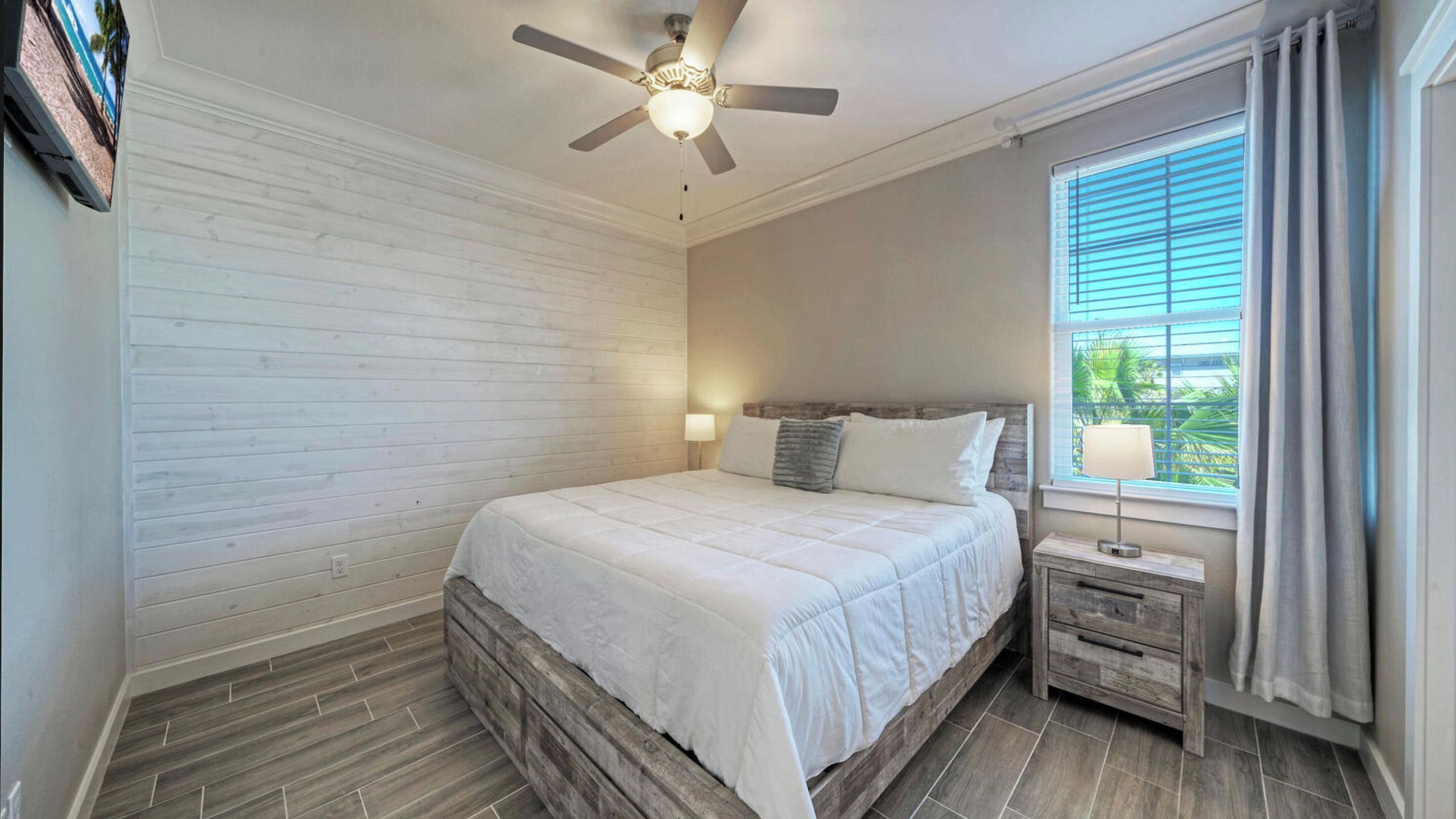 Un dormitorio con cama, ventilador de techo, mesita de noche, lámpara y ventana con cortinas.