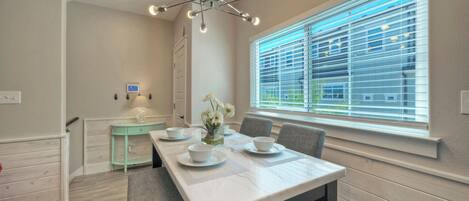 Un comedor con una mesa preparada para cuatro, una lámpara moderna, una ventana con persianas y una pequeña mesita de noche con lámpara.