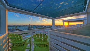 A balcony with two green chairs, a view of the sunset over the water, and a building in the background.