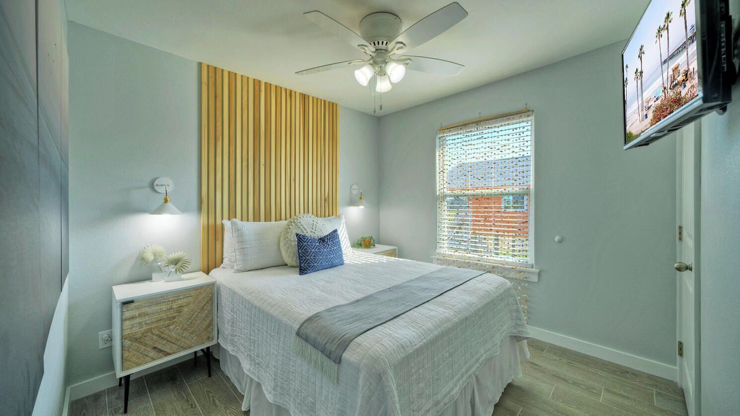 A bedroom with a bed, nightstands, a ceiling fan, a window with blinds, and a TV mounted on the wall.