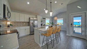 A modern kitchen with white cabinets, a central island with bar stools, a stainless steel refrigerator, and a chandelier. - Outdoor Lounge w/ Bar Swing Seats, Near Nature Preserve Blutopia by Avantstay (Port Aransas)