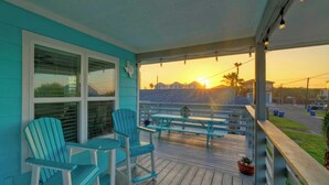 Un porche con dos sillas Adirondack y una mesa, con vista a una playa y un atardecer.