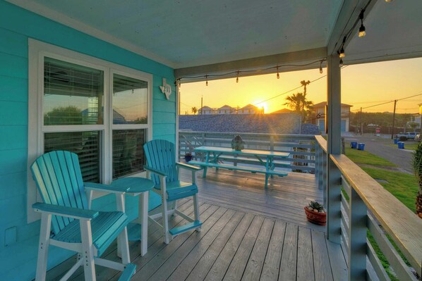 Une véranda avec deux fauteuils Adirondack et une table, donnant sur une plage où l’on peut admirer le coucher de soleil.