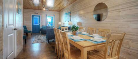 A wooden interior with a dining table set for six, chairs, a blue door, and a ceiling fan.