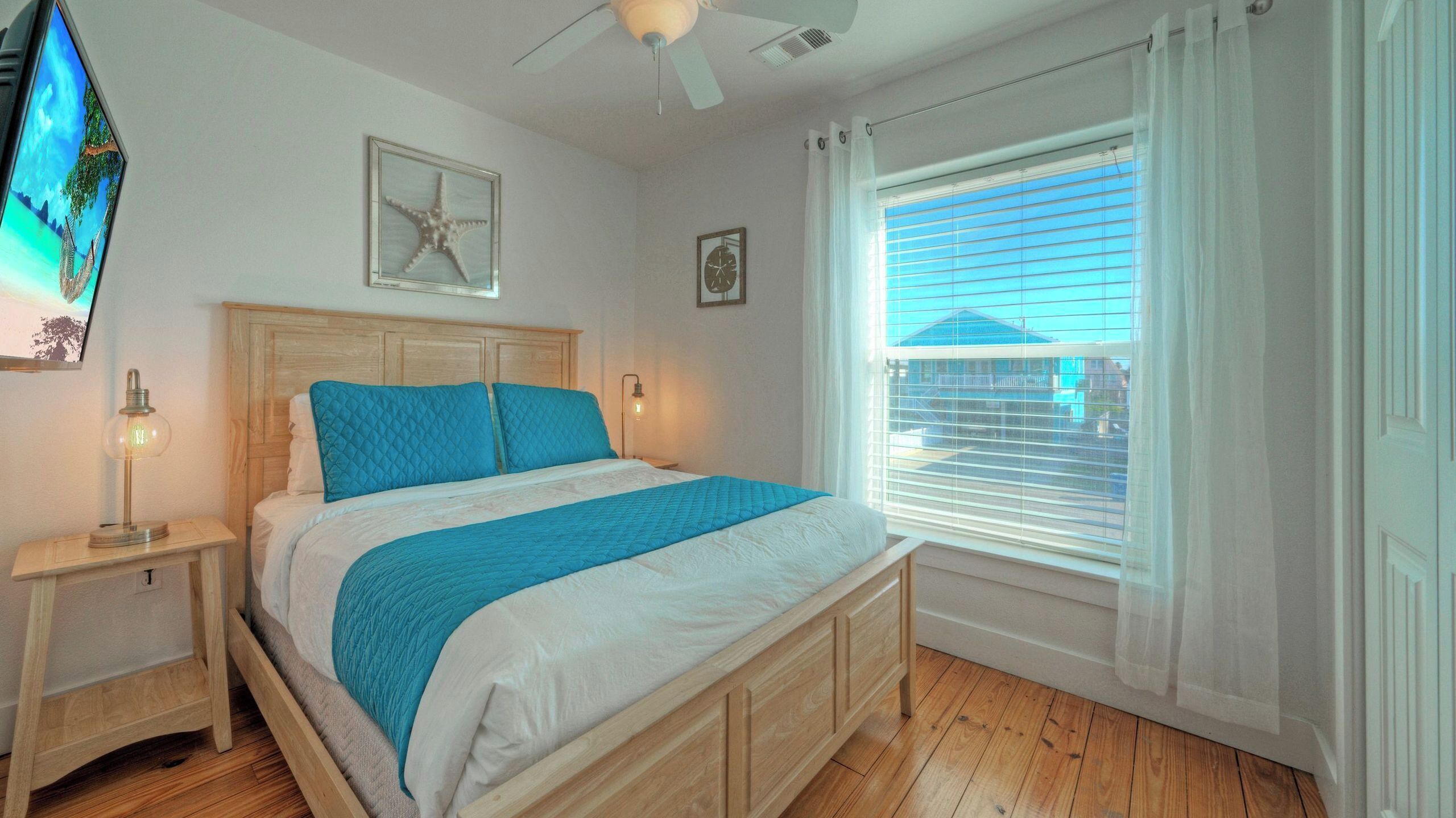 A bedroom with a bed, wooden headboard, bedside tables, a ceiling fan, a window with blinds, and a wall-mounted TV.