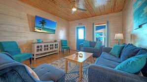 A cozy room with a wooden ceiling, a ceiling fan, a TV on the wall, a cabinet, two sofas, a coffee table, and a window. - Pool, Patio, Blocks to the Beach Golf Cart Zone Daiquiri Daze by Avantstay (Port Aransas)