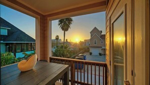 Un balcon en bois avec une table et des chaises, donnant sur un quartier résidentiel avec des maisons et un coucher de soleil.