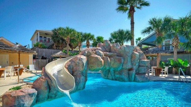 Une piscine avec un toboggan, des palmiers et une terrasse en bois avec des chaises.