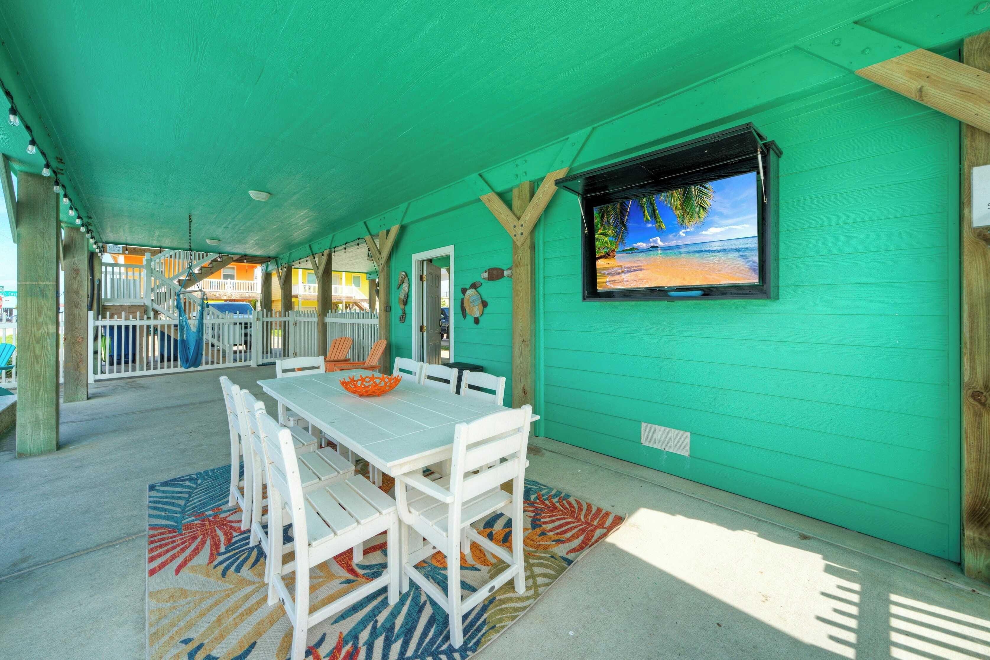 A covered outdoor dining area with a table and chairs, a television screen displaying a beach scene, and a view of a building with a white fence.