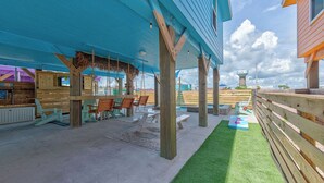 A covered outdoor seating area with wooden chairs and tables, a thatched roof, and a view of a water tower in the background. - Totally Beachin by Avantstay Private Heated Pool, Hot Tub + Fire Pit! (Port Aransas)