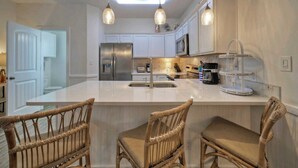 A modern kitchen with white cabinetry, a stainless steel refrigerator, a double sink, and a breakfast bar with wicker chairs. - 2 Blocks to the Beach, Patio, Resort Pool w/ Slide Tranquil Tides by Avantstay (Port Aransas)