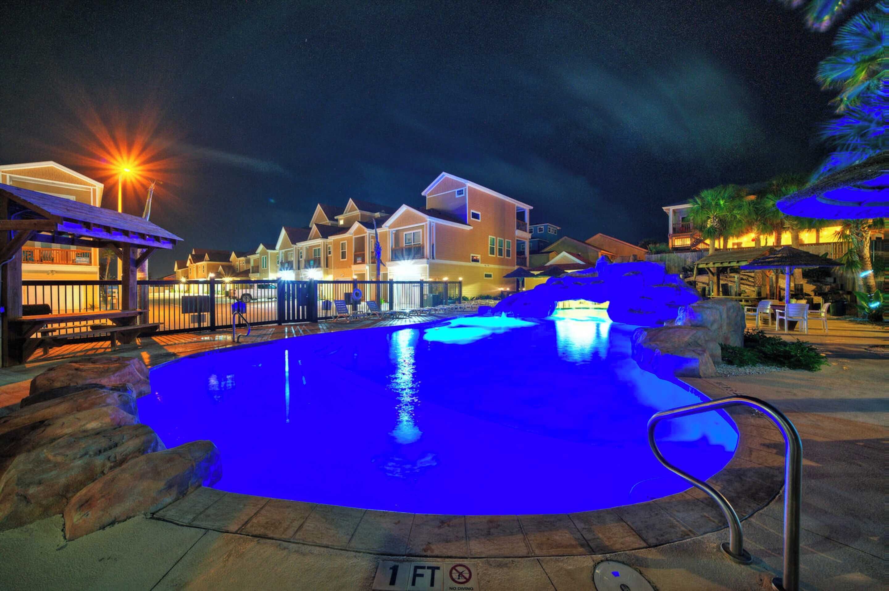 A swimming pool with a 1 FT no diving sign, surrounded by a deck with outdoor seating and a night-time illuminated background.