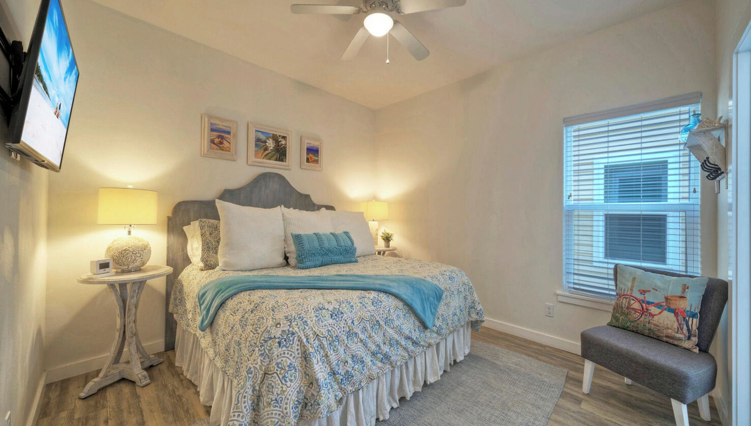 A bedroom with a bed, two bedside lamps, a ceiling fan, a TV mounted on the wall, and a window with blinds.