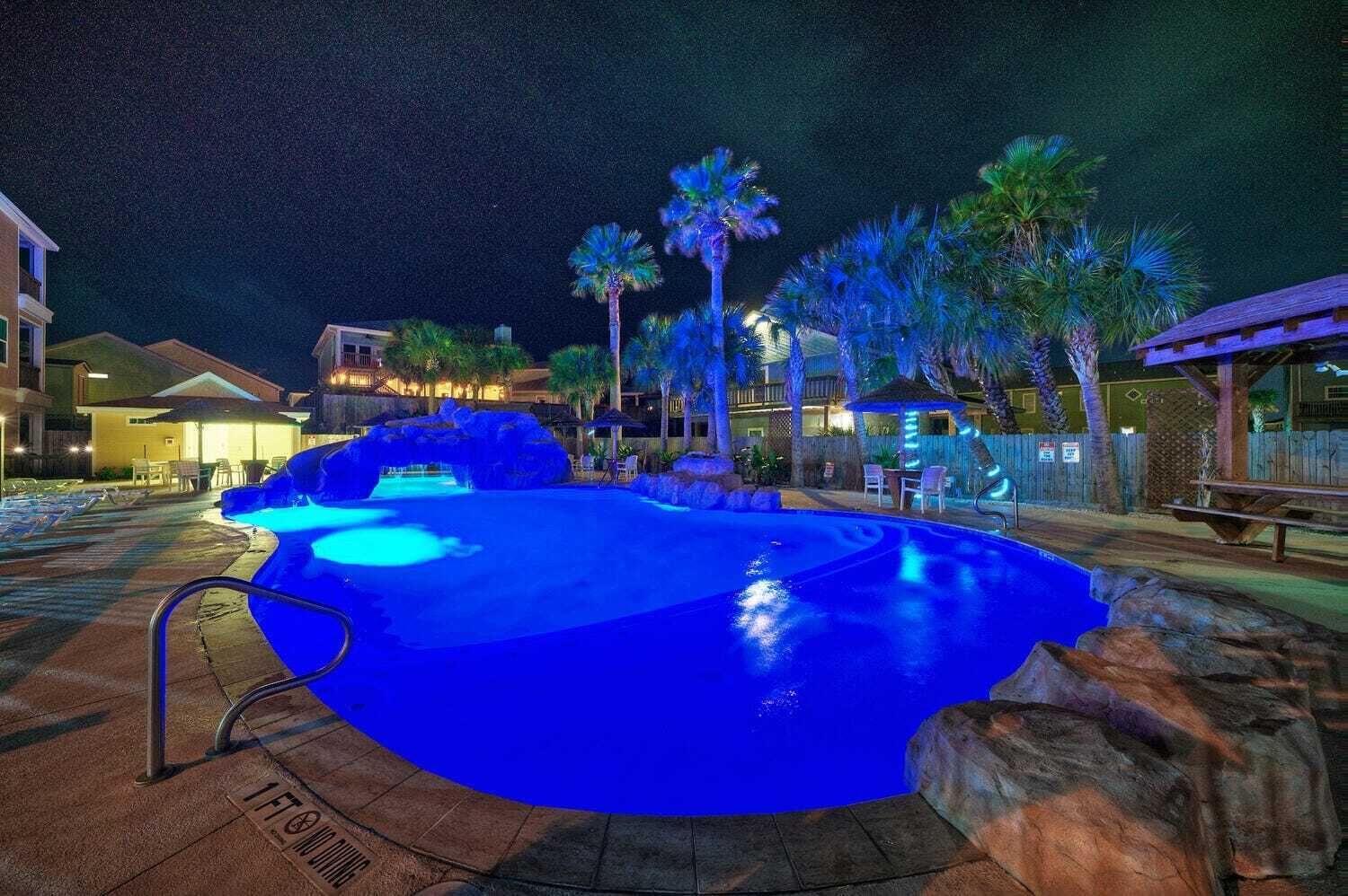 A nighttime swimming pool with a blue light, surrounded by palm trees and a wooden deck.