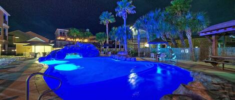 A nighttime swimming pool with a blue light, surrounded by palm trees and a wooden deck.