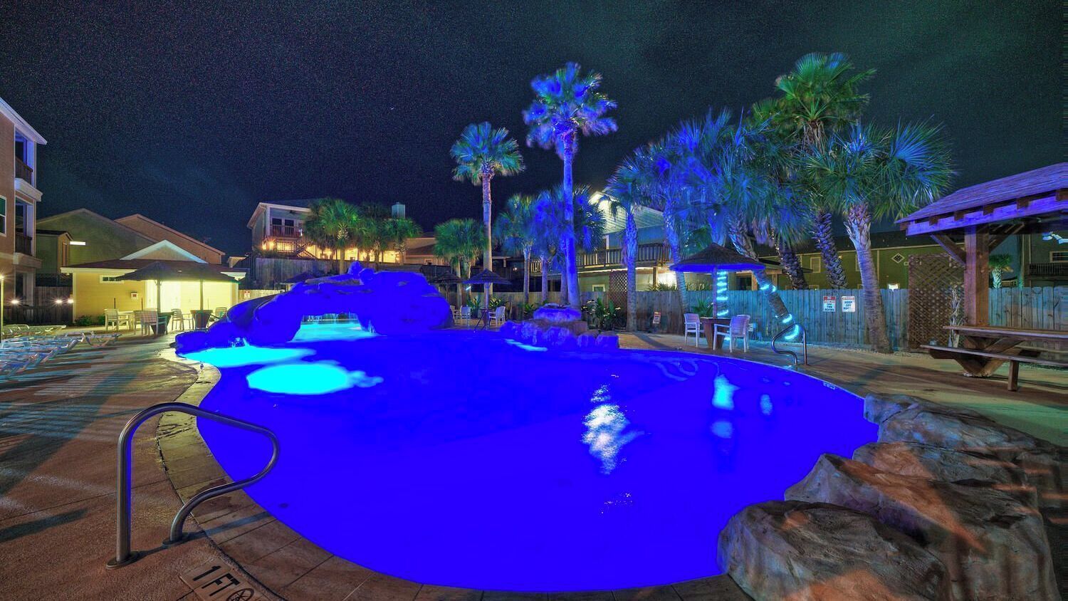 A nighttime swimming pool with blue lighting, palm trees, and a wooden deck.