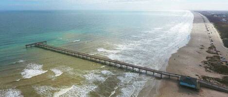 Vista aérea de una playa con un muelle largo que se adentra en el océano, una orilla arenosa y algunos edificios cerca de la línea de costa.