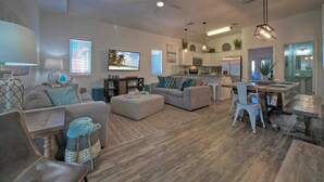 A spacious living room with a sofa, ottoman, and dining table. There is a kitchen area with cabinets and a refrigerator. - Isla View by Avantstay Resort Pool, Near Beach (Port Aransas)