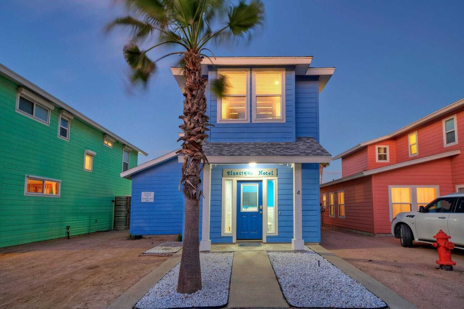 A hotel with a blue facade, a palm tree in front, and a white car parked nearby.