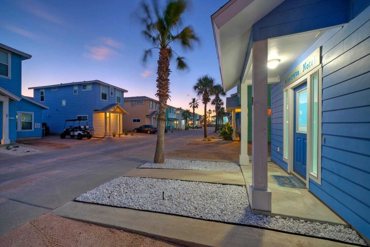 Un hotel con fachada azul, zona de estacionamiento y palmeras.