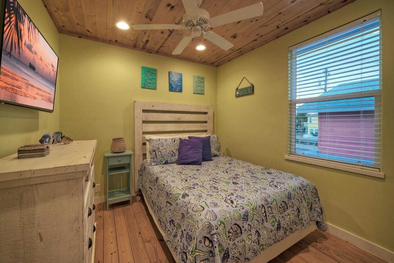 A bedroom with a wooden ceiling, a bed with patterned bedding, a nightstand, a small table with decorative items, and a window with blinds.