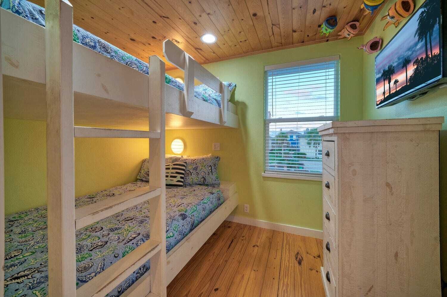 Een kamer met stapelbedden, een raam met blinds, een houten kast en een aan de muur bevestigde televisie.