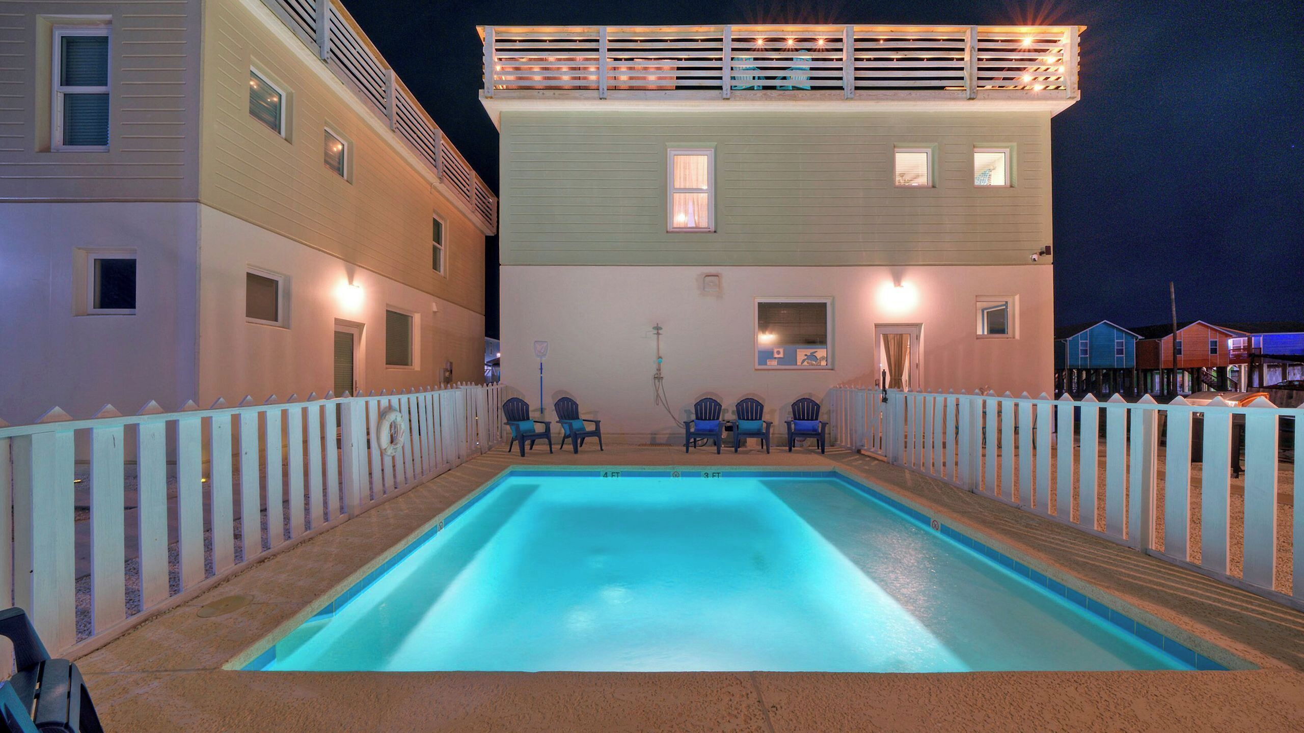 A swimming pool with chairs, a white fence, and buildings in the background.