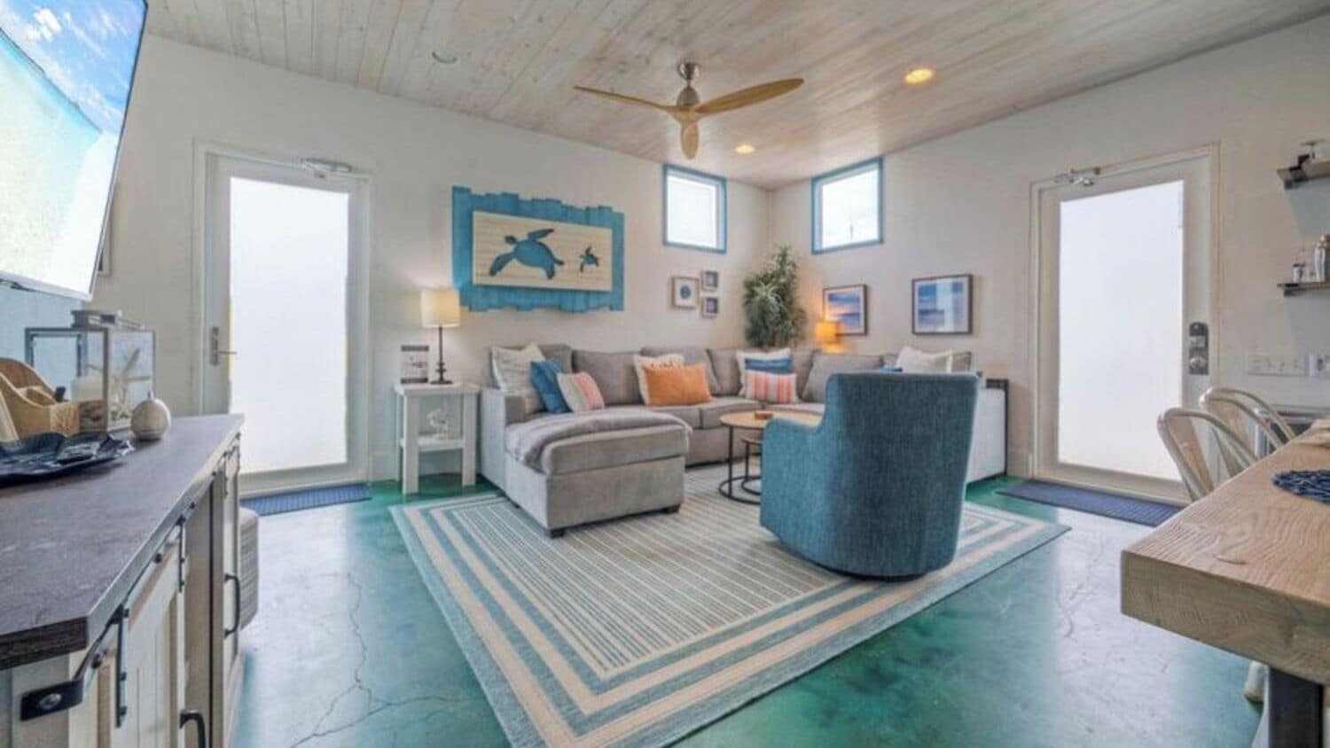 A spacious living area with a sofa, armchair, and a dining table. There is a ceiling fan, a large window, and a wall-mounted artwork.