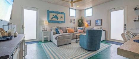 A spacious living area with a sofa, armchair, and a dining table. There is a ceiling fan, a large window, and a wall-mounted artwork.