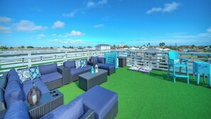 Una terraza en la azotea con mobiliario exterior, una valla de madera y vistas al océano y edificios.
