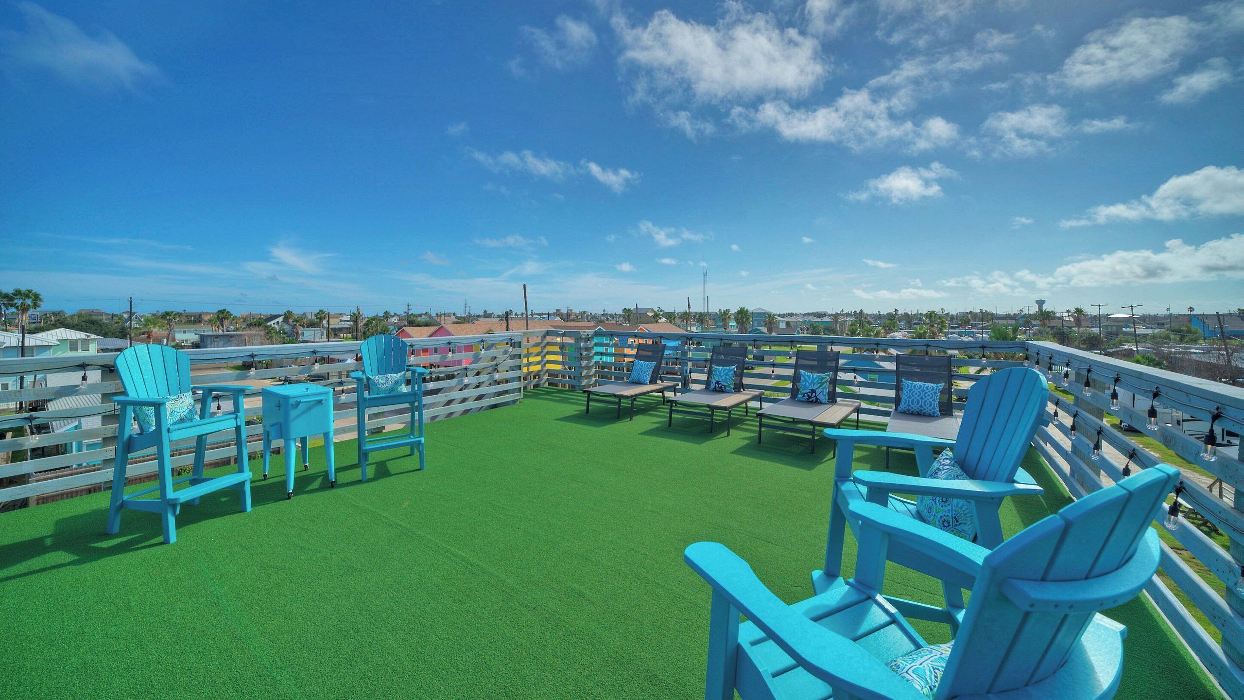 A rooftop terrace with blue Adirondack chairs, a green artificial turf, and a view of a coastal area.