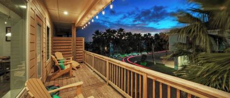 A balcony with wooden chairs and a railing overlooking a road and palm trees.