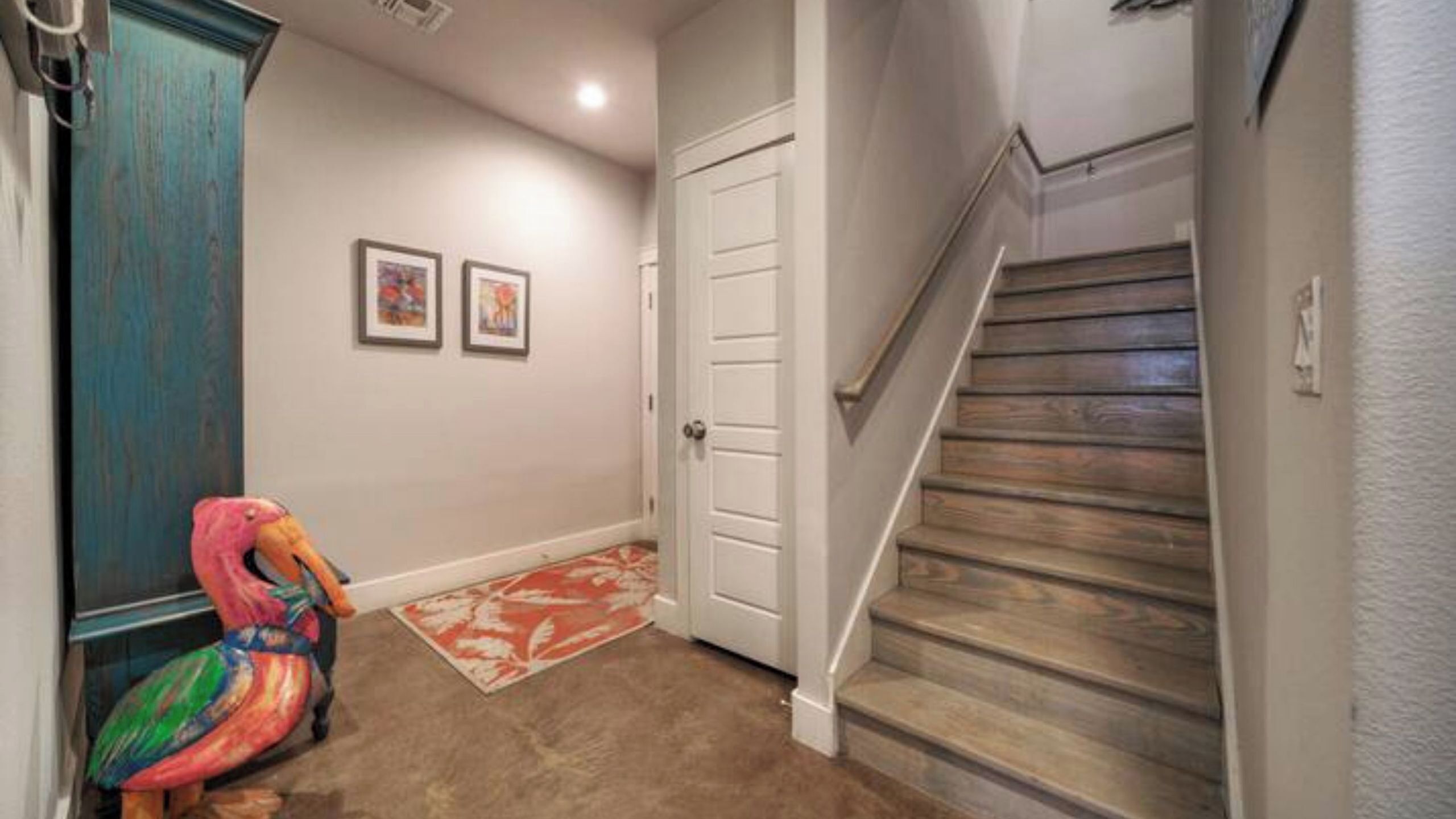 A hallway with a wooden staircase, a door, and a colorful flamingo sculpture.