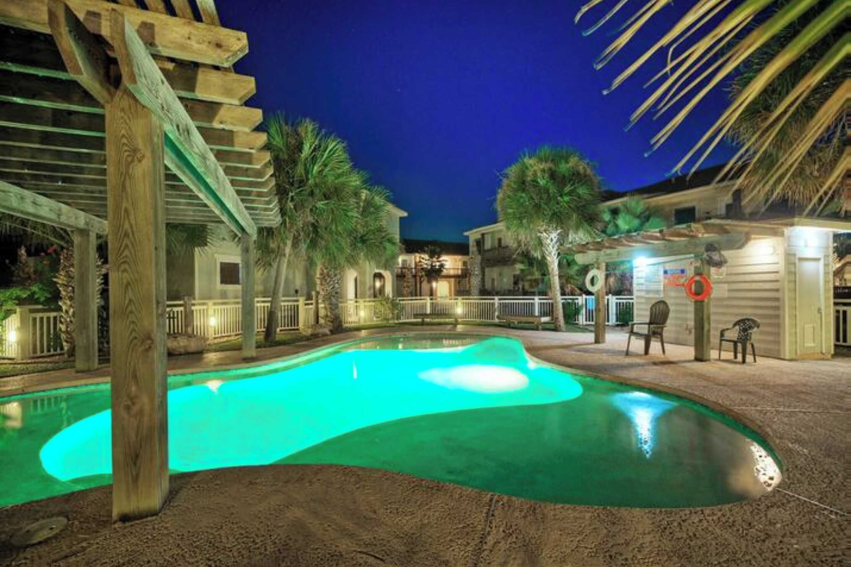 A swimming pool with a wooden pergola structure, surrounded by palm trees and a building with a balcony.