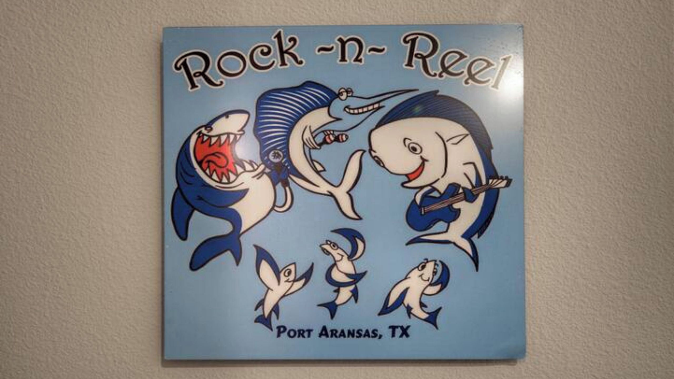 Un autocollant mural représentant un requin de dessin animé et des poissons jouant de la guitare, avec les inscriptions « Rock-n-Reel » et « Port Aransas, TX ».