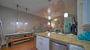 Une cuisine dotée d’un plan de travail en granit, de meubles blancs et d’un lave-vaisselle en acier inoxydable. On y trouve également un banc en bois et une table à manger avec des chaises.