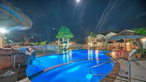 Un espace piscine nocturne doté d’une piscine à eau claire, d’un poste de surveillance pour sauveteur et d’une petite cascade décorative.