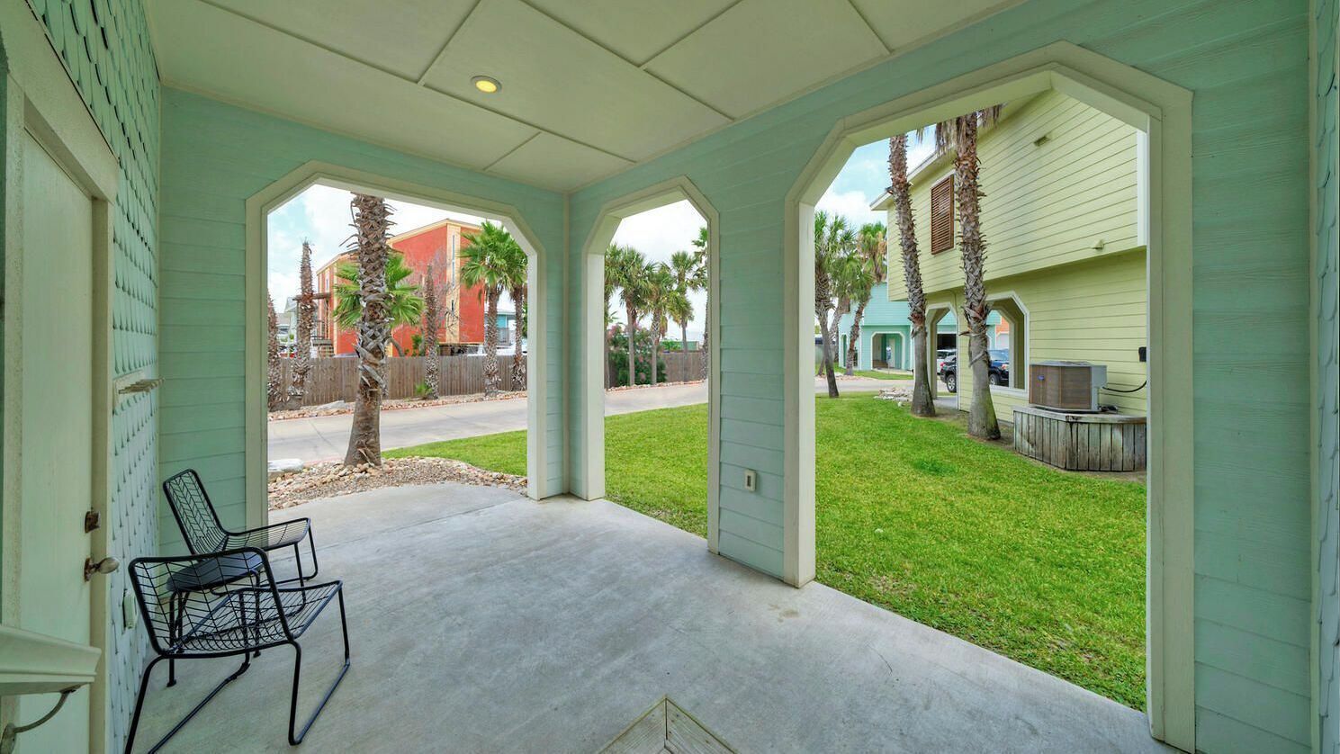 A covered porch with a concrete floor, two metal chairs, and a view of a green lawn and neighboring houses.