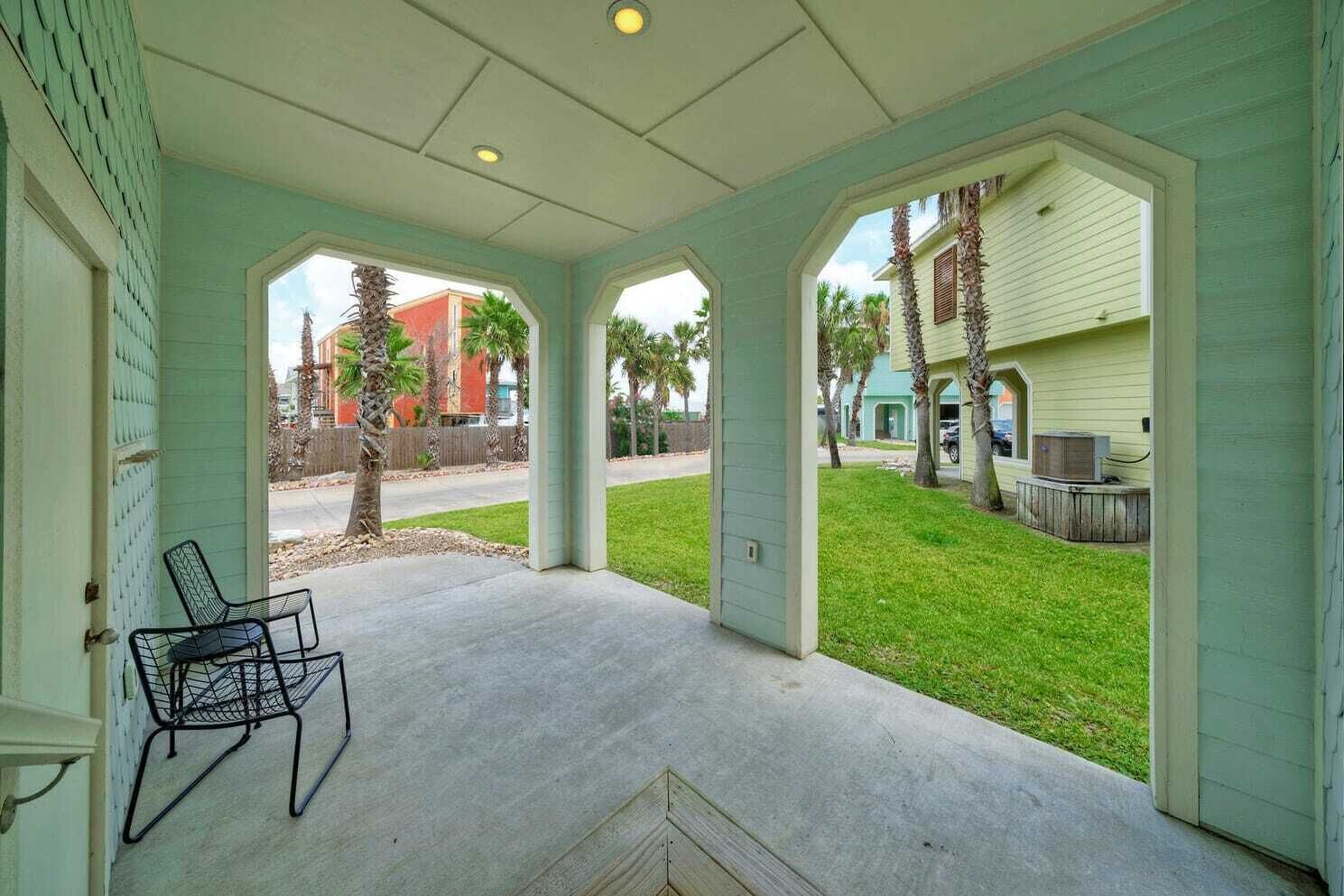 A covered porch with a concrete floor and metal chairs, overlooking a well-maintained lawn and neighboring houses.