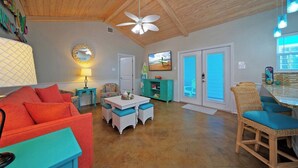 A spacious living area with a ceiling fan, a television, a dining table with chairs, and a wicker chair. - Coastal Vibe by Avantstay Sunset at the Pool and Playscape (Port Aransas)