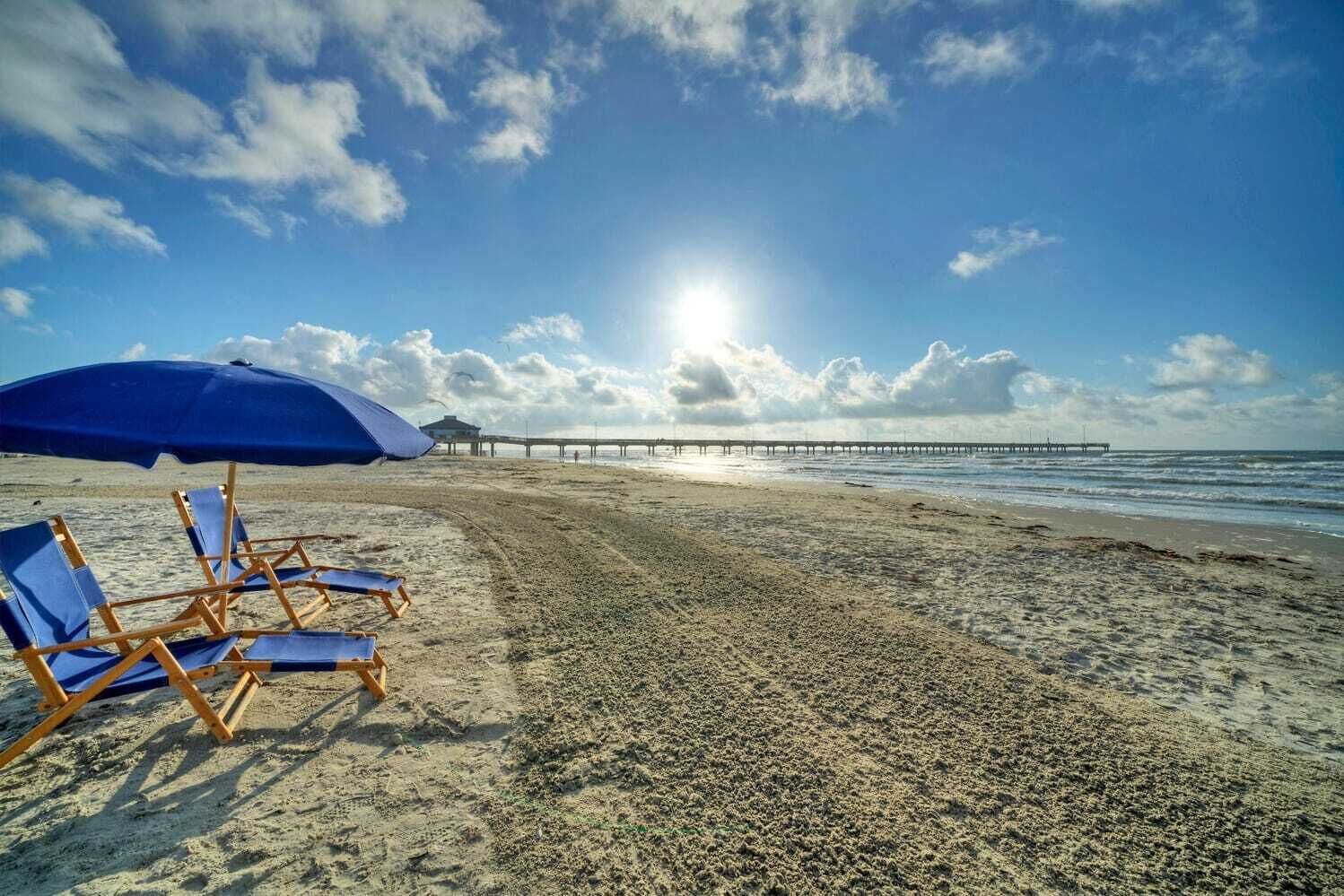 Una spiaggia con sedie a sdraio blu e un ombrellone, un molo che si estende nell'acqua, e un cielo sereno.