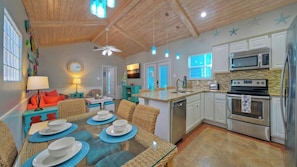 A modern kitchen with white cabinets, stainless steel appliances, and a dining area with wicker chairs and blue placemats. - Coastal Vibe by Avantstay Sunset at the Pool and Playscape (Port Aransas)