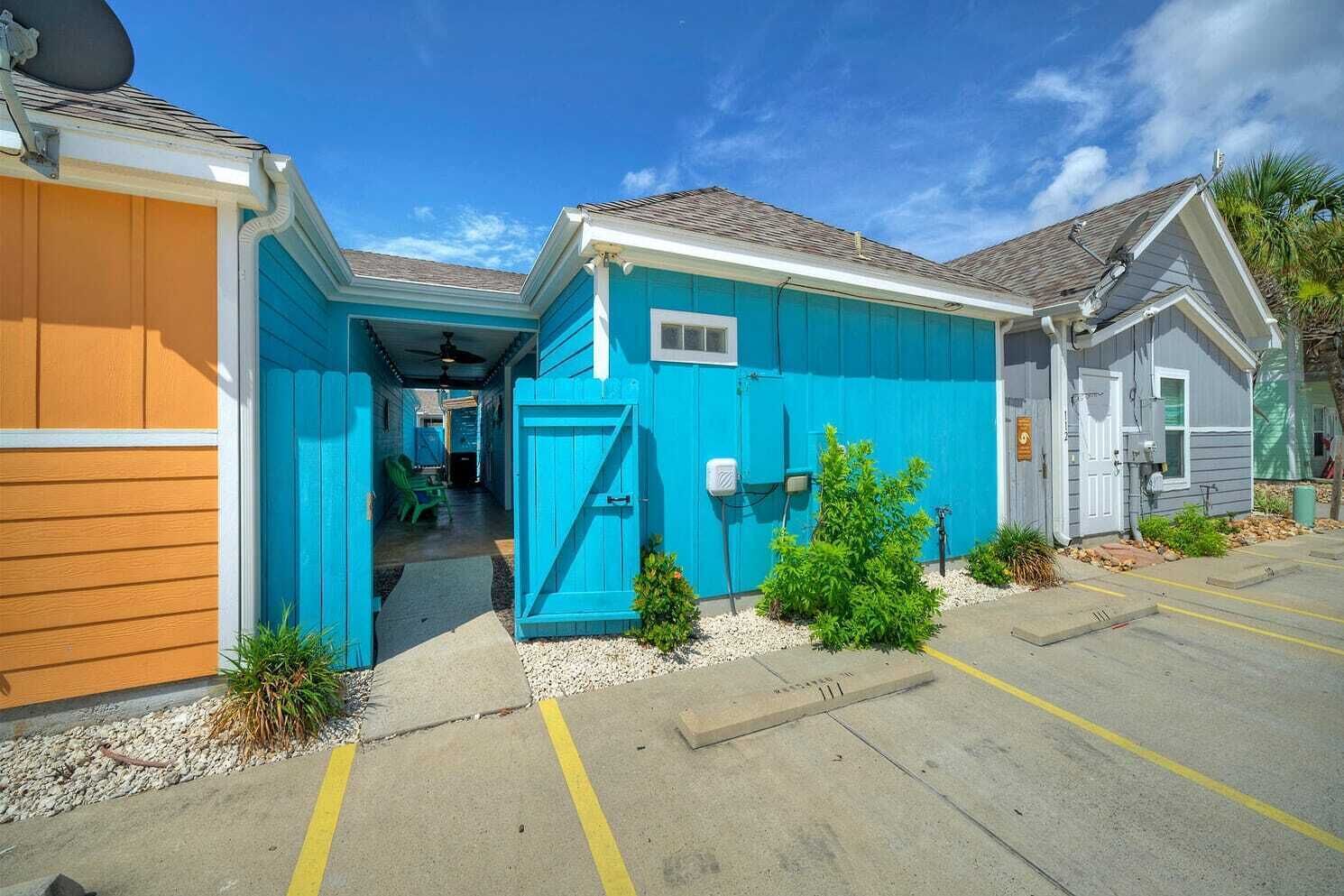 A row of small, single-story buildings with a blue exterior, a covered walkway, and a parking area with yellow lines.