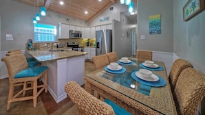 A modern kitchen with white cabinets, a central island, and a dining area with wicker chairs and a glass table set for four. - Coastal Vibe by Avantstay Sunset at the Pool and Playscape (Port Aransas)