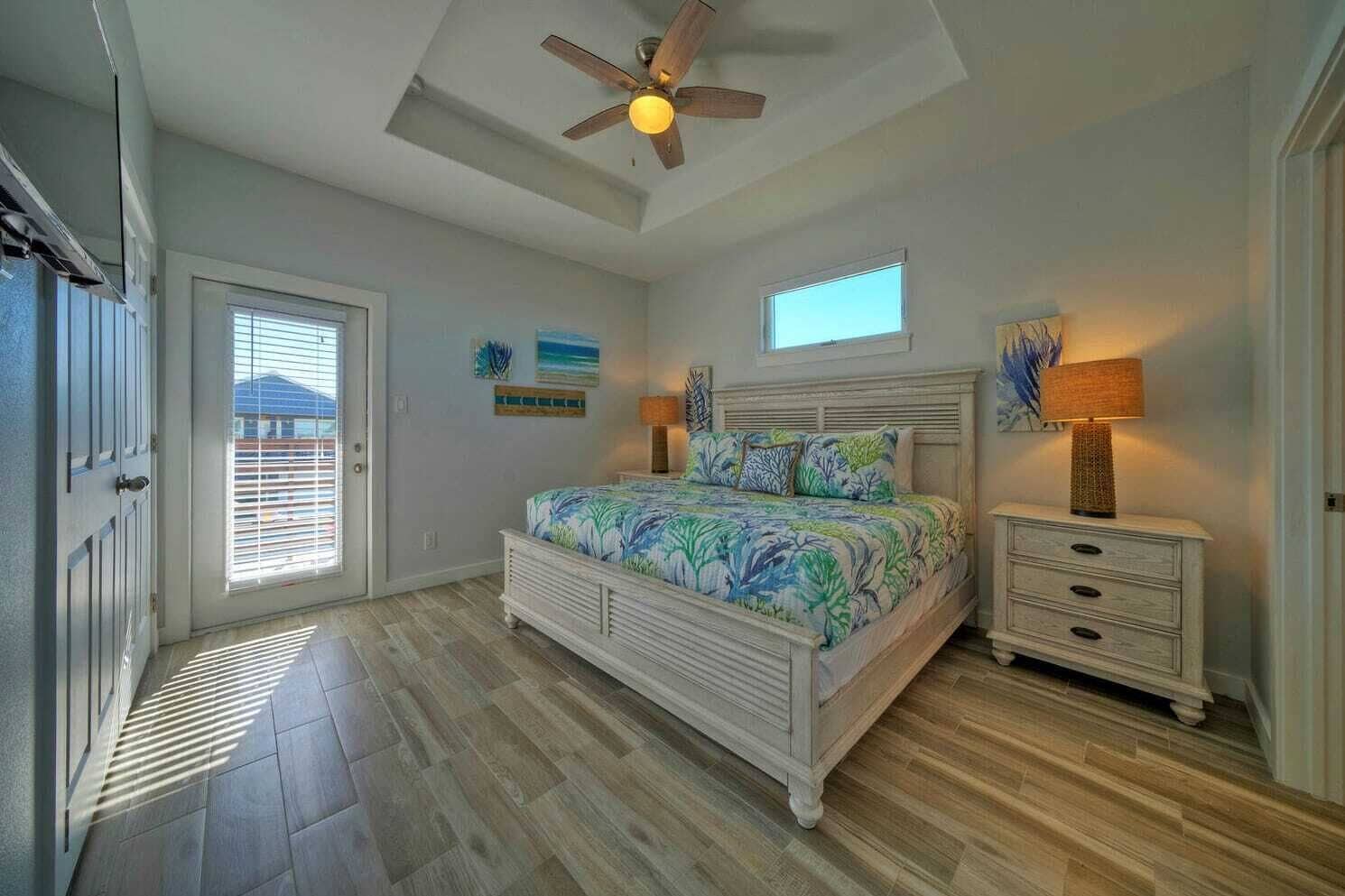 Quarto com cama, mesa de cabeceira, ventilador de teto, janela e porta.