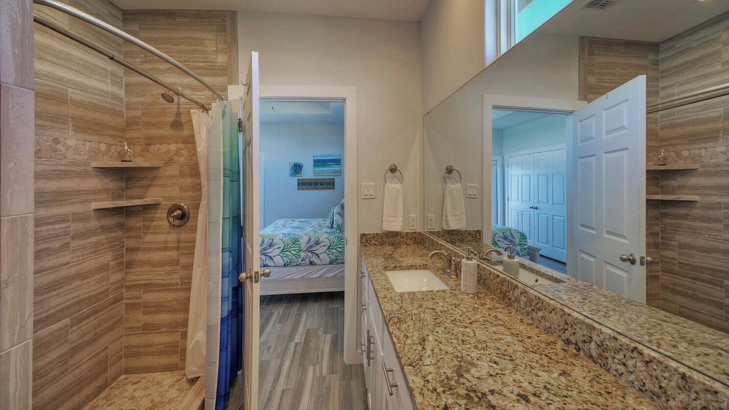 A bathroom with a shower, a sink, a mirror, and a door leading to another room.